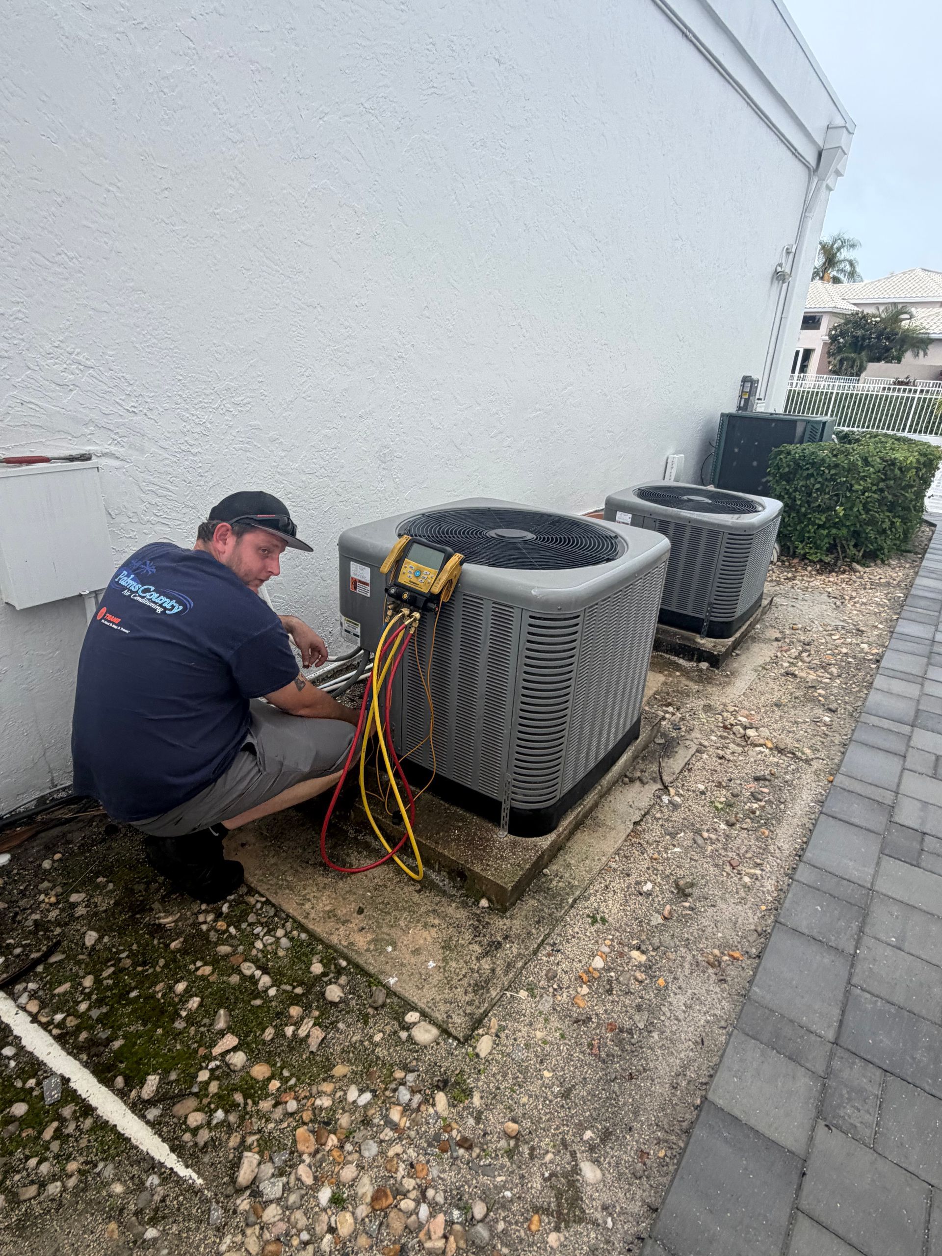 Technician servicing outdoor HVAC units beside a building wall, with tools and hoses on the ground.