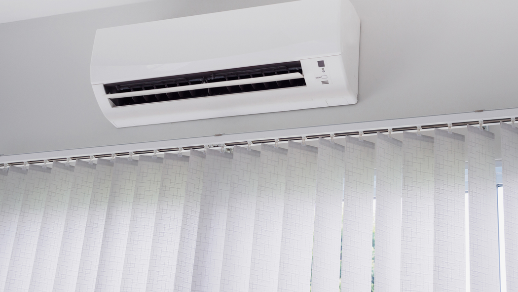 White wall-mounted air conditioner above white vertical blinds in a bright room