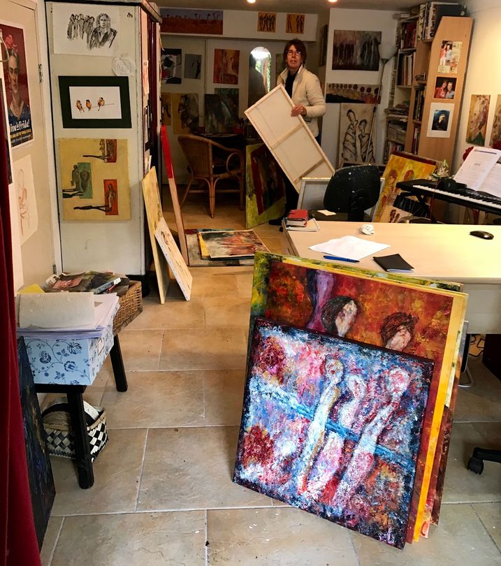 Kunststudio met schilderijen en materialen; op de achtergrond is een persoon te zien die een ingelijst doek draagt.