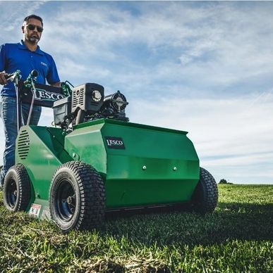 Man operating a green lawn aerator on a grassy field under a blue sky