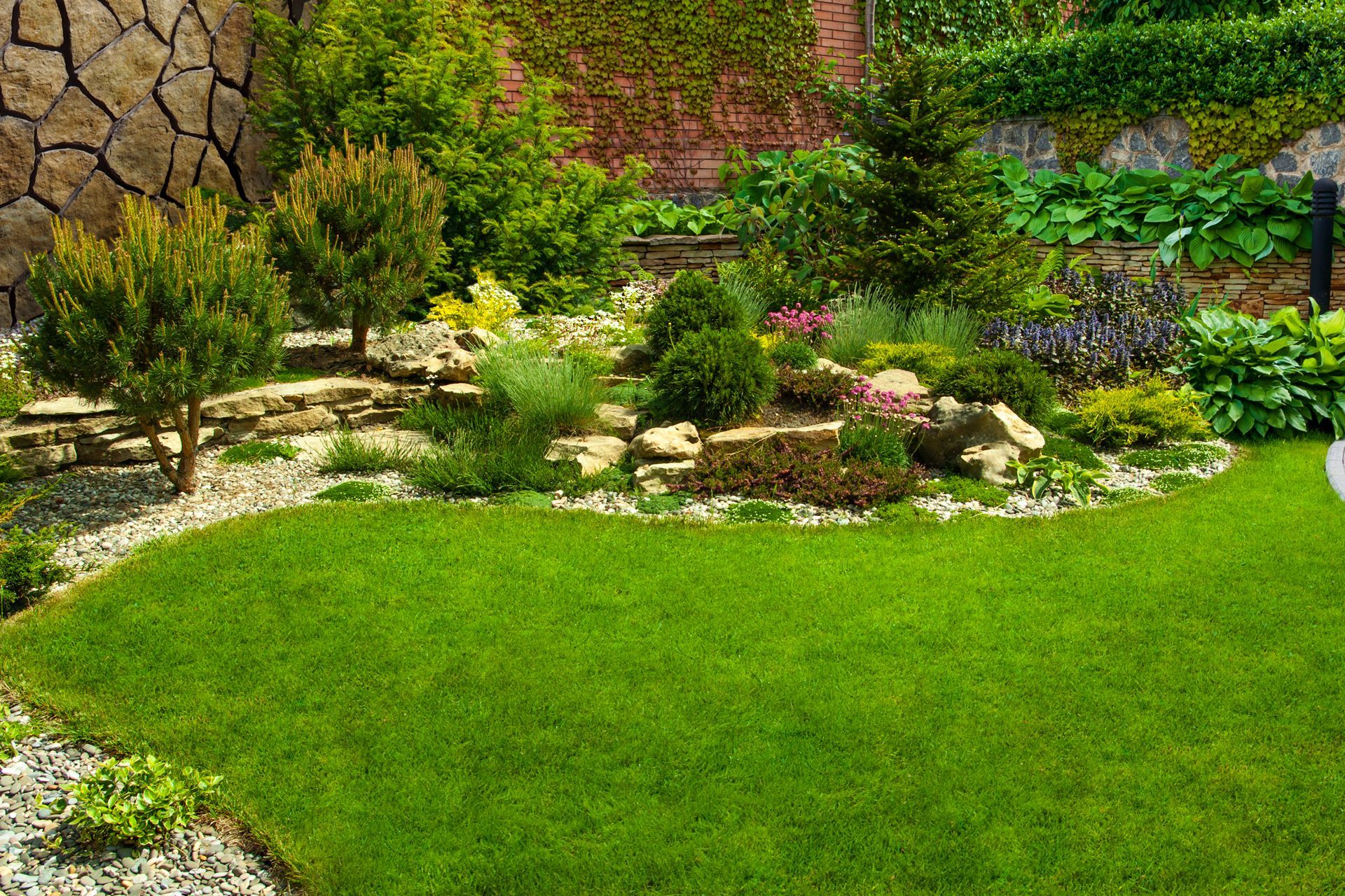 Lush green lawn curving to a garden of shrubs, rocks, and trees against a stone wall backdrop