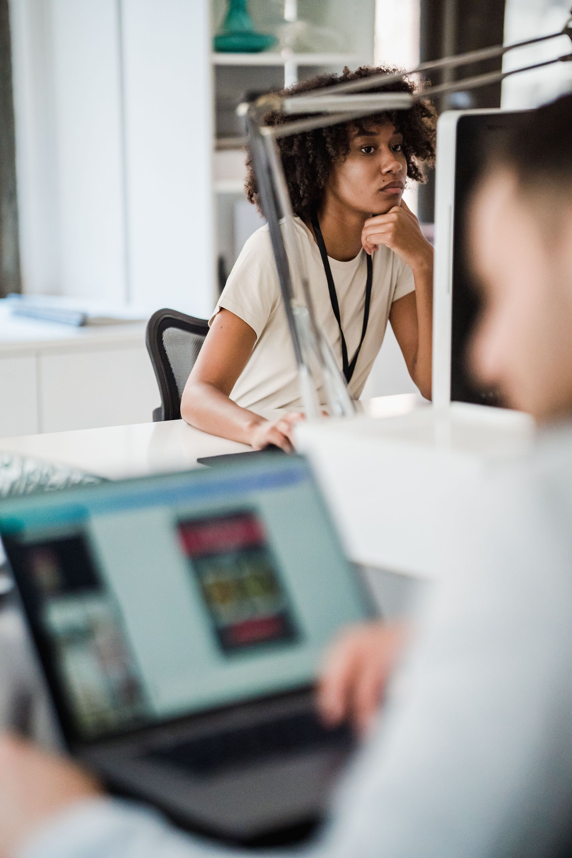 Person wearing a white shirt sits at a laptop in a bright office, looking thoughtful through a divider
