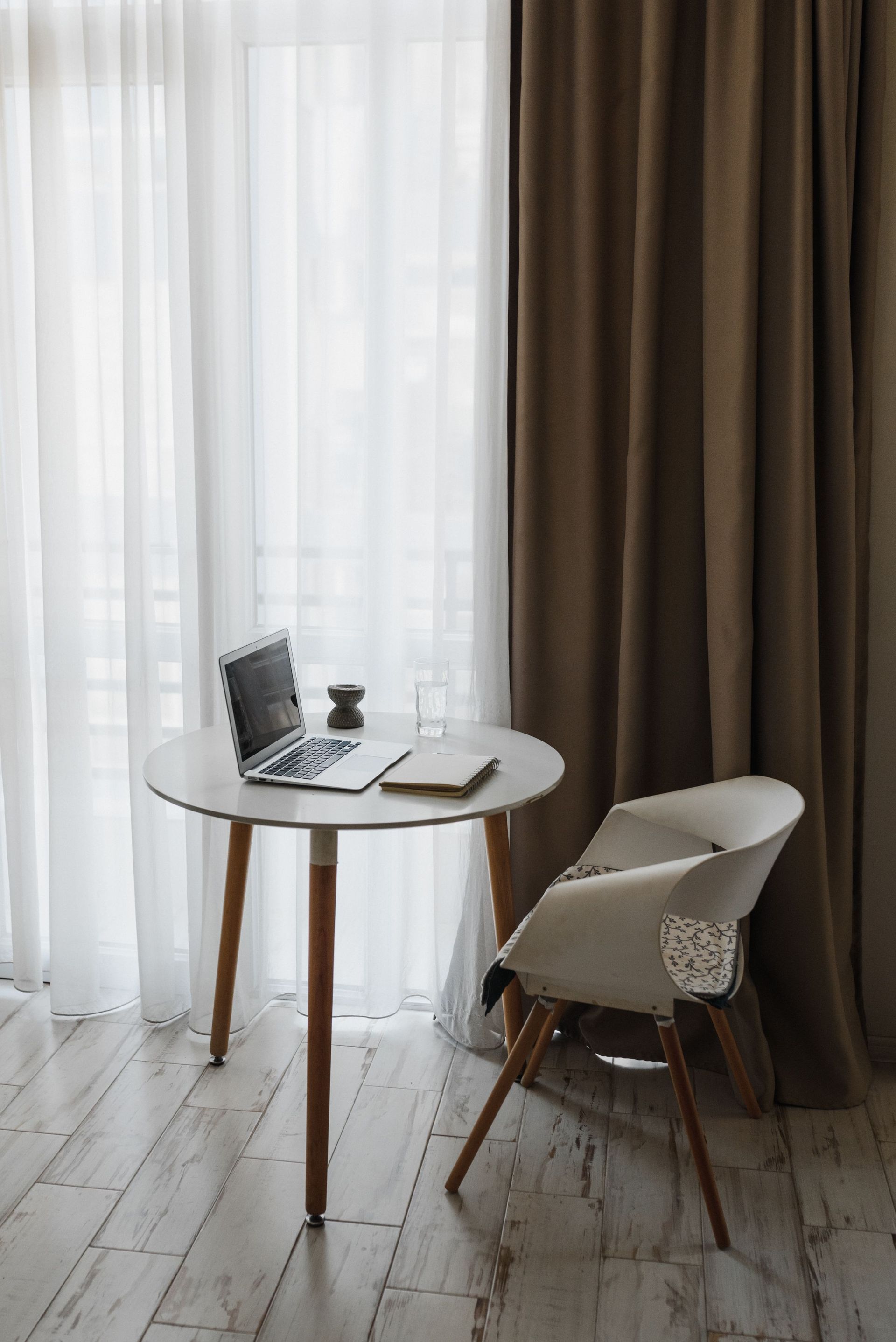 Small round table with laptop and coffee beside a white chair by sheer curtains and brown drapes