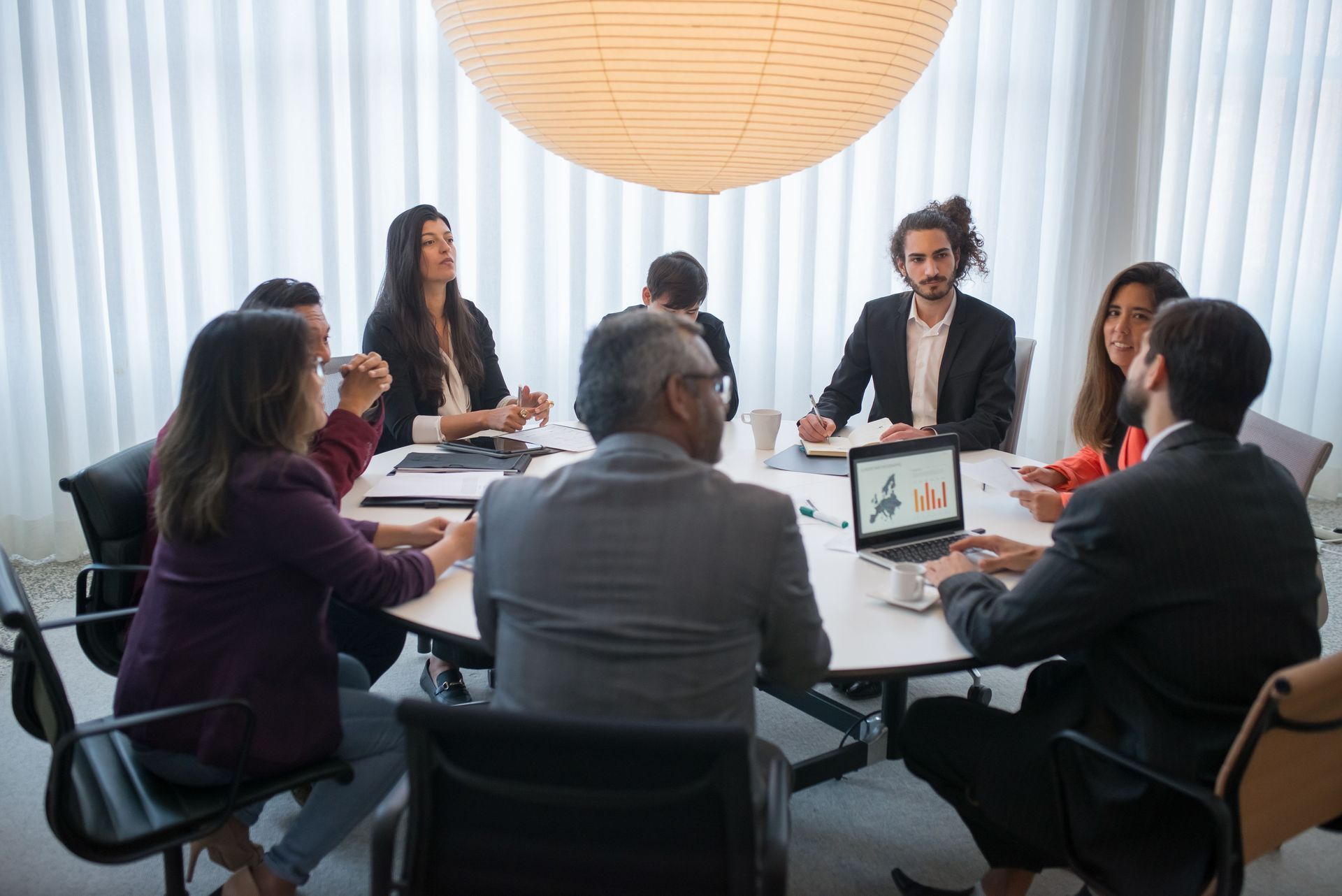 Team meeting around a conference table in a bright office, with a laptop and laptop screen visible.
