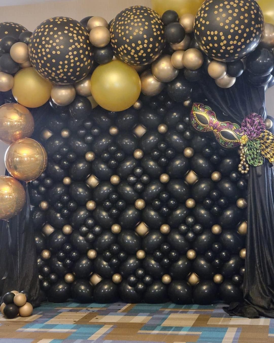 A wall filled with black and gold balloons and a mask.