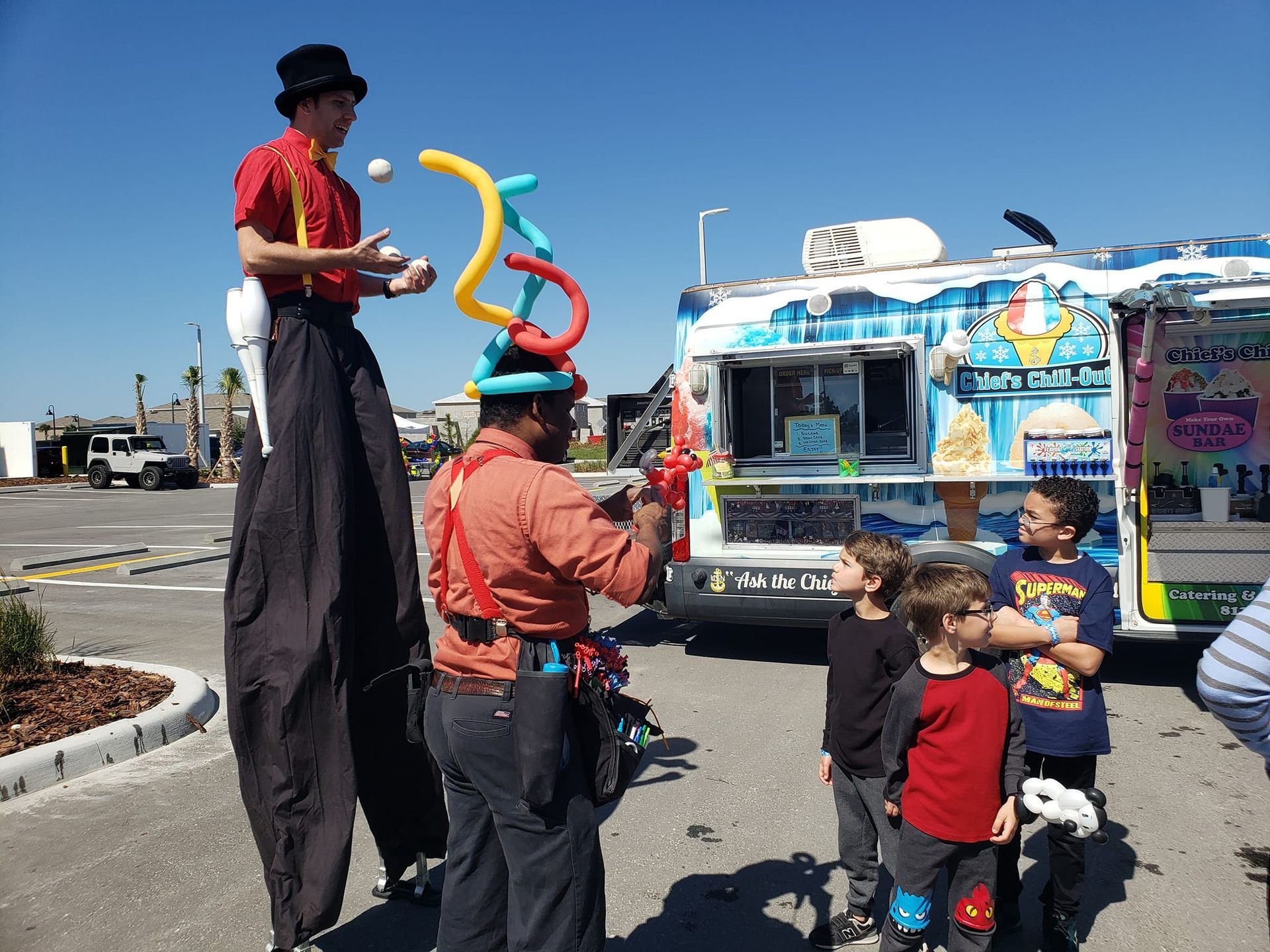 A man on stilts is playing with balloons in front of an ice cream truck.