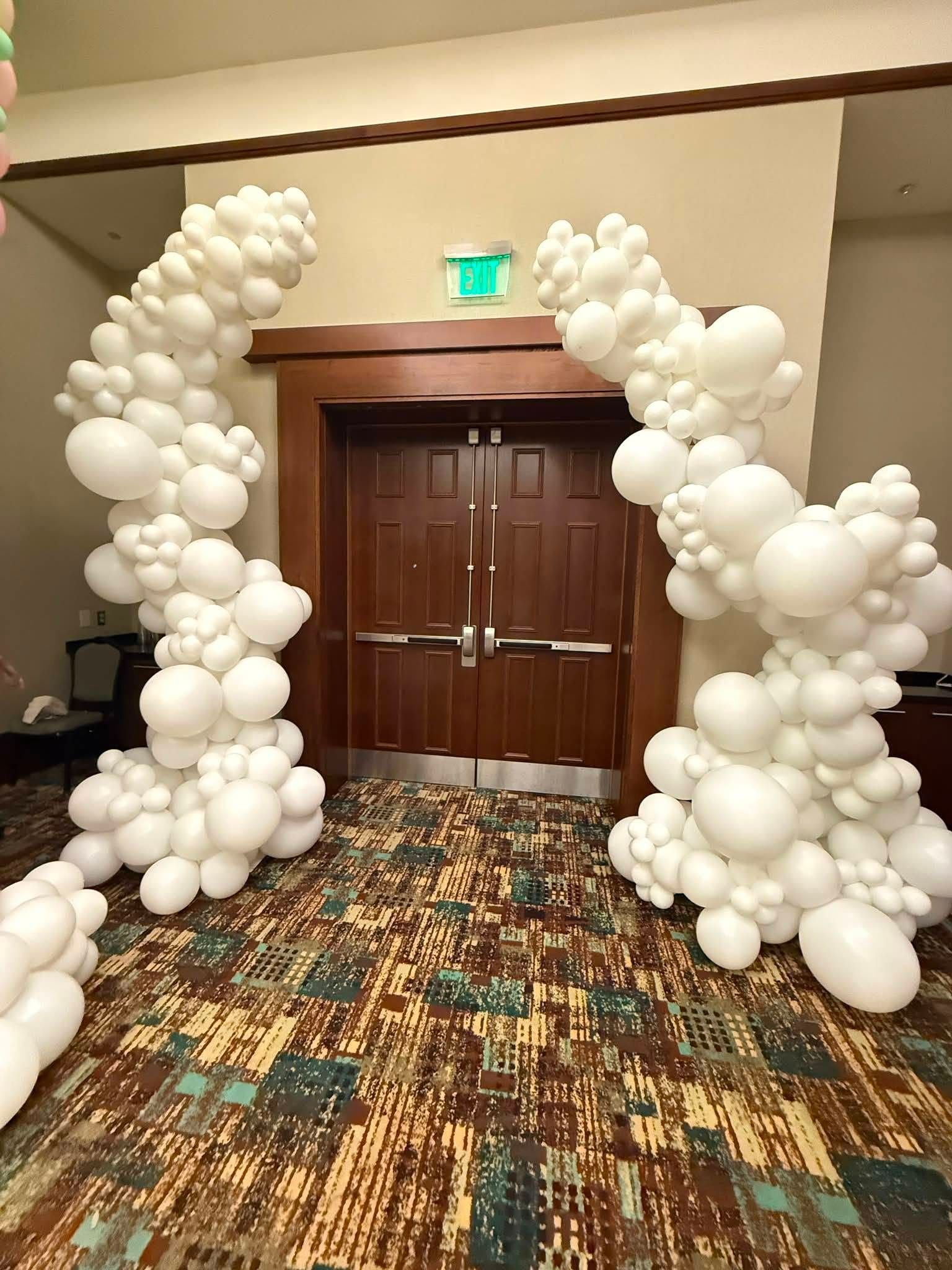White balloon arch framing wooden double doors in a hotel hallway with patterned carpet