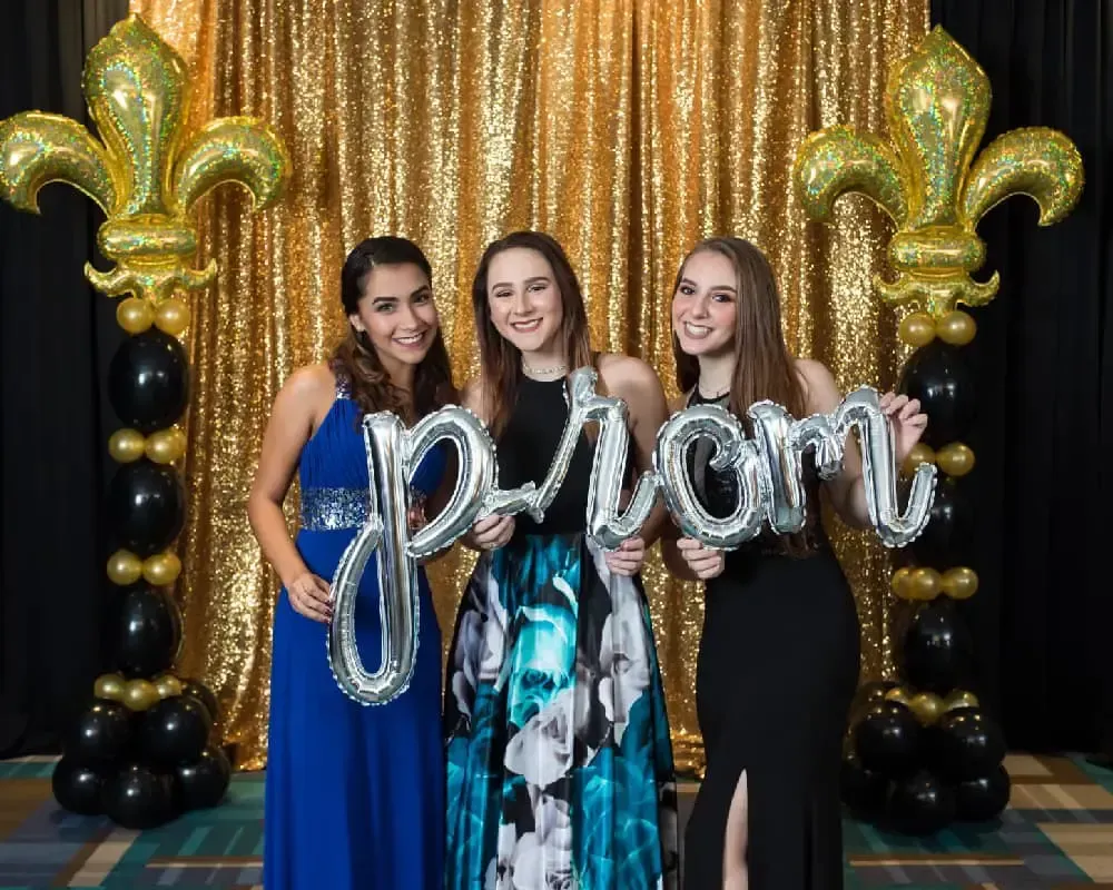 Three girls are standing in front of a gold curtain holding balloons that spell out the word prom.