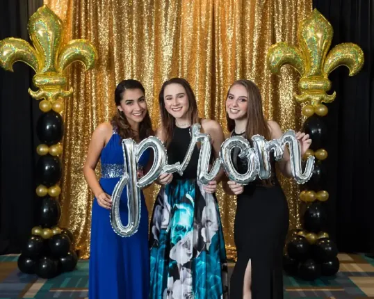 Three girls are standing in front of a gold curtain holding balloons that spell out the word prom.