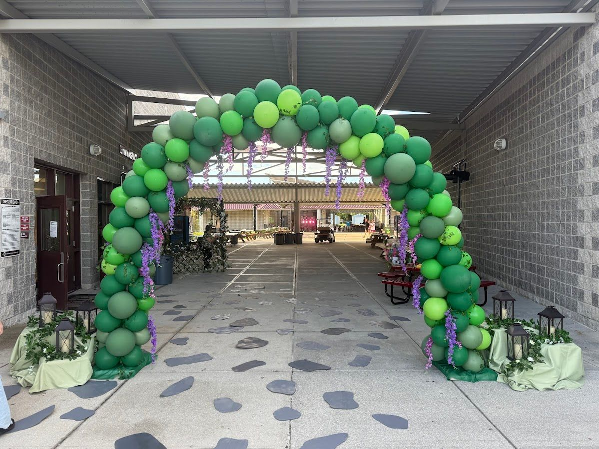 A large green balloon arch decorated with purple accents, standing at the entrance to a stone-walled outdoor courtyard.