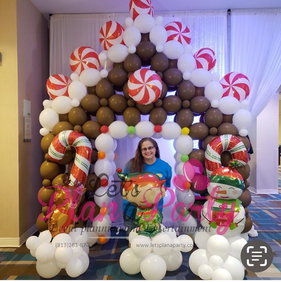 A person stands in a large gingerbread house balloon arch decorated with candy-themed balloons, a candy cane, and an elf.