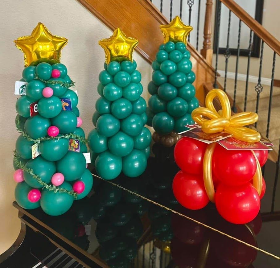 Three green balloon Christmas trees topped with gold stars and a red balloon gift box sit on a black piano.