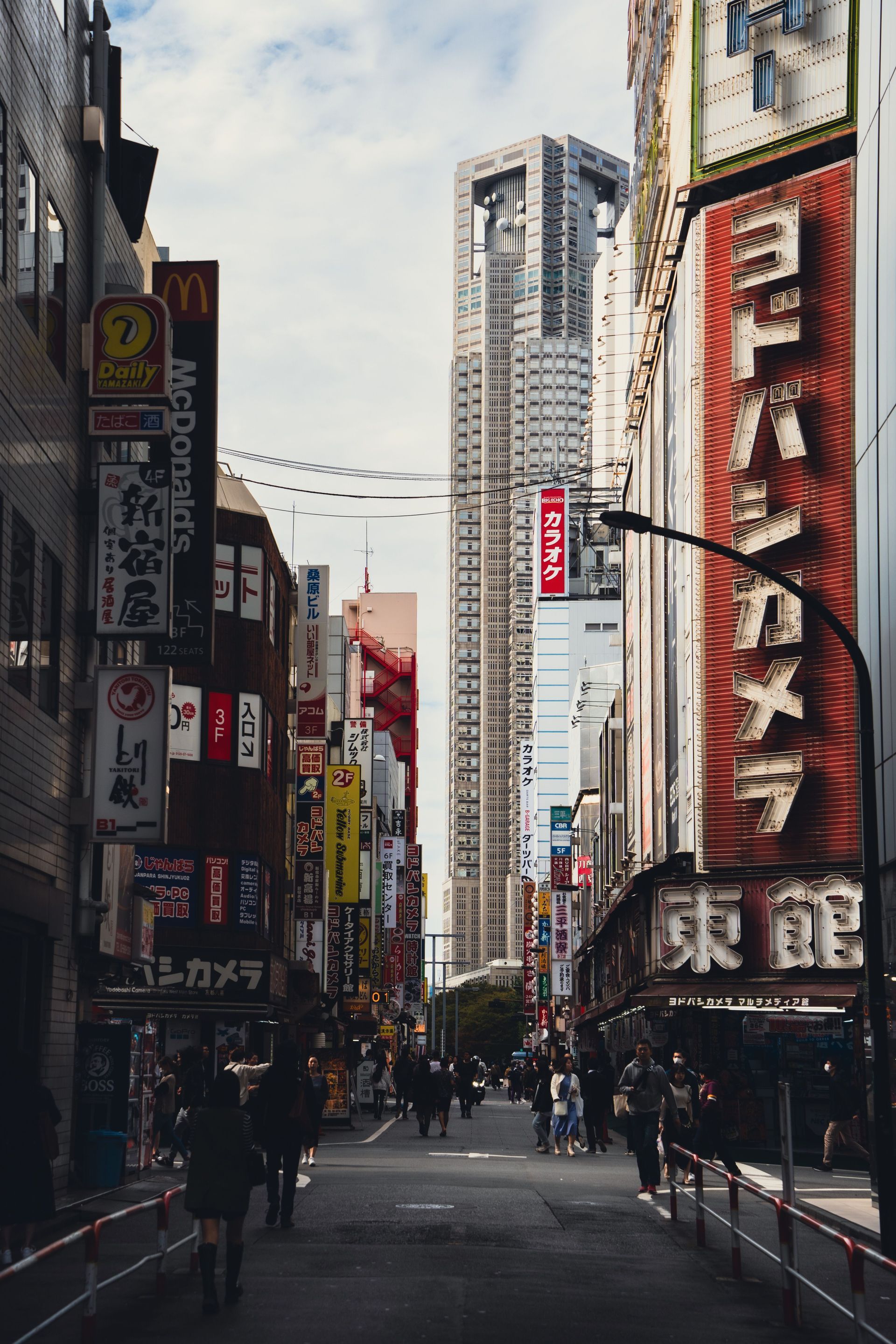 Eine enge Straße in Tokio mit hohen Gebäuden, Leuchtreklamen und Fußgängern in der Ferne.
