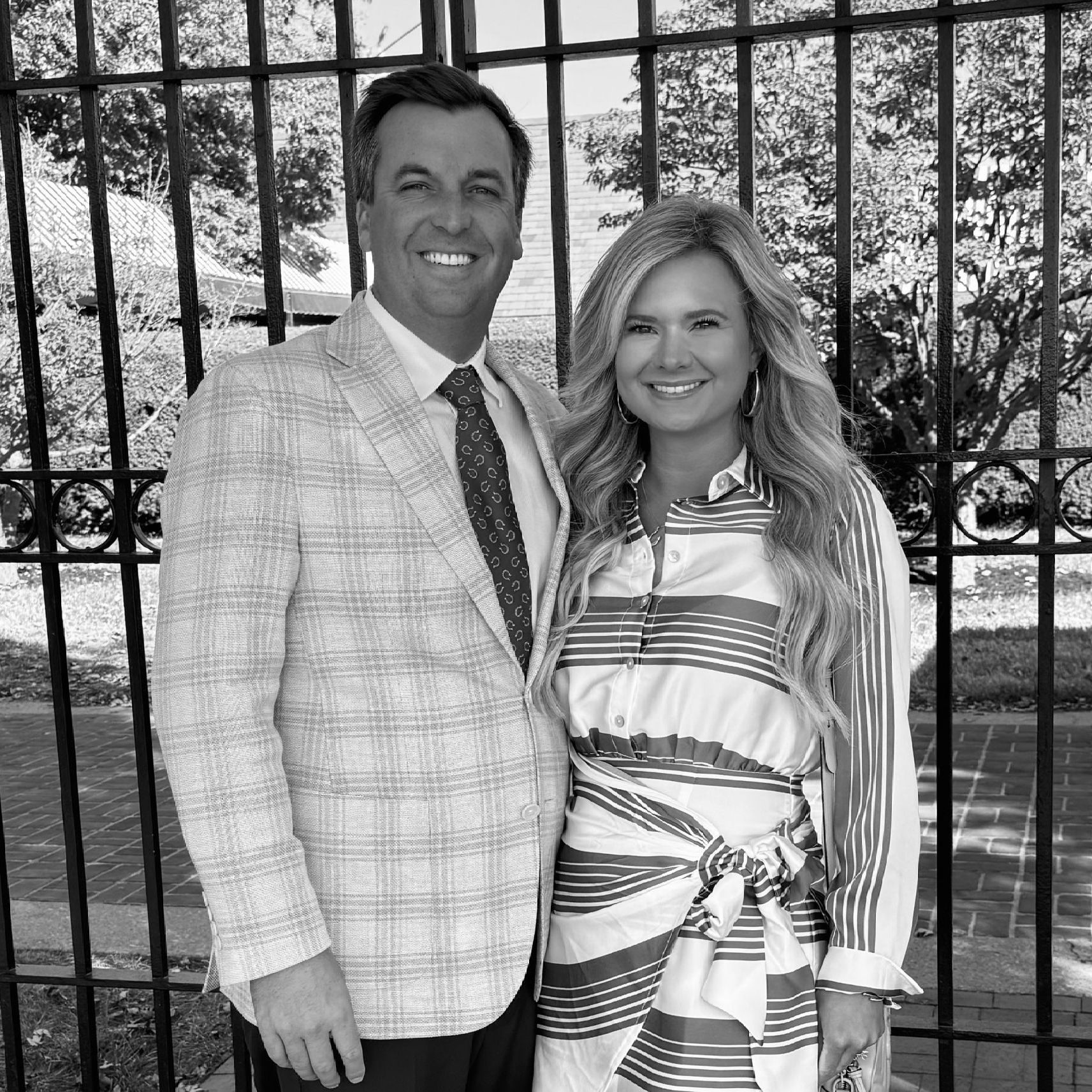 A couple stands smiling in front of a black iron gate outdoors, wearing formal attire.