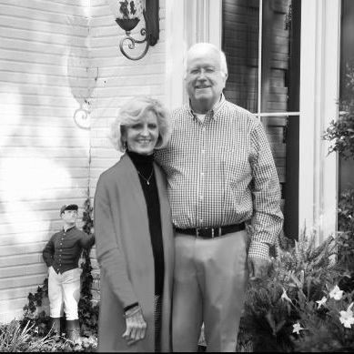A man and woman stand smiling side-by-side on a porch, next to a small lawn jockey statue near a building exterior.