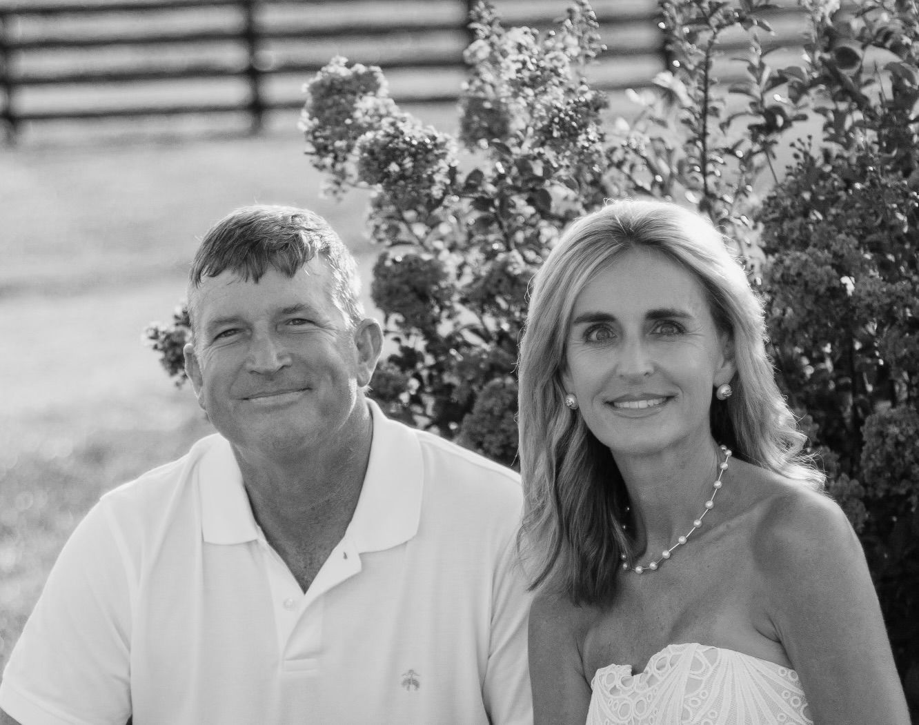 A smiling couple sits together outdoors in front of a leafy bush and a fence, captured in a black and white photograph.