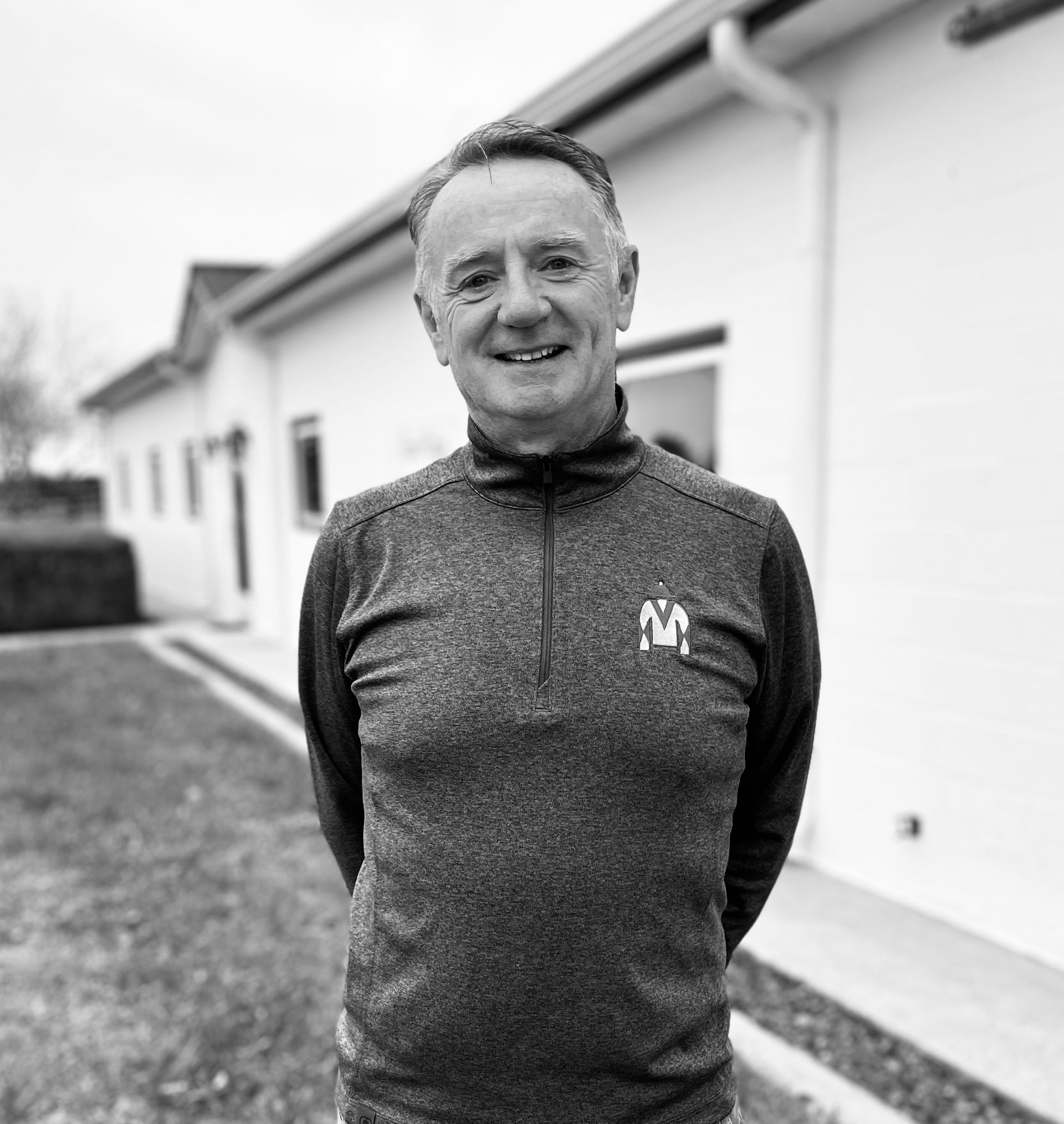 A person stands smiling in a quarter-zip pullover in front of a white building on a clear day.