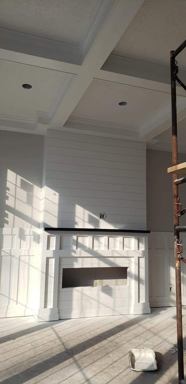 Sunlit modern living room with white paneled fireplace wall and tall windows casting shadows on the floor