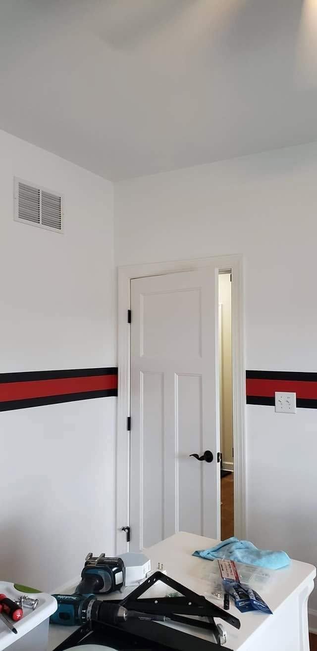 Small white bedroom with a red-and-black wall stripe, open door, and cluttered bed in the foreground