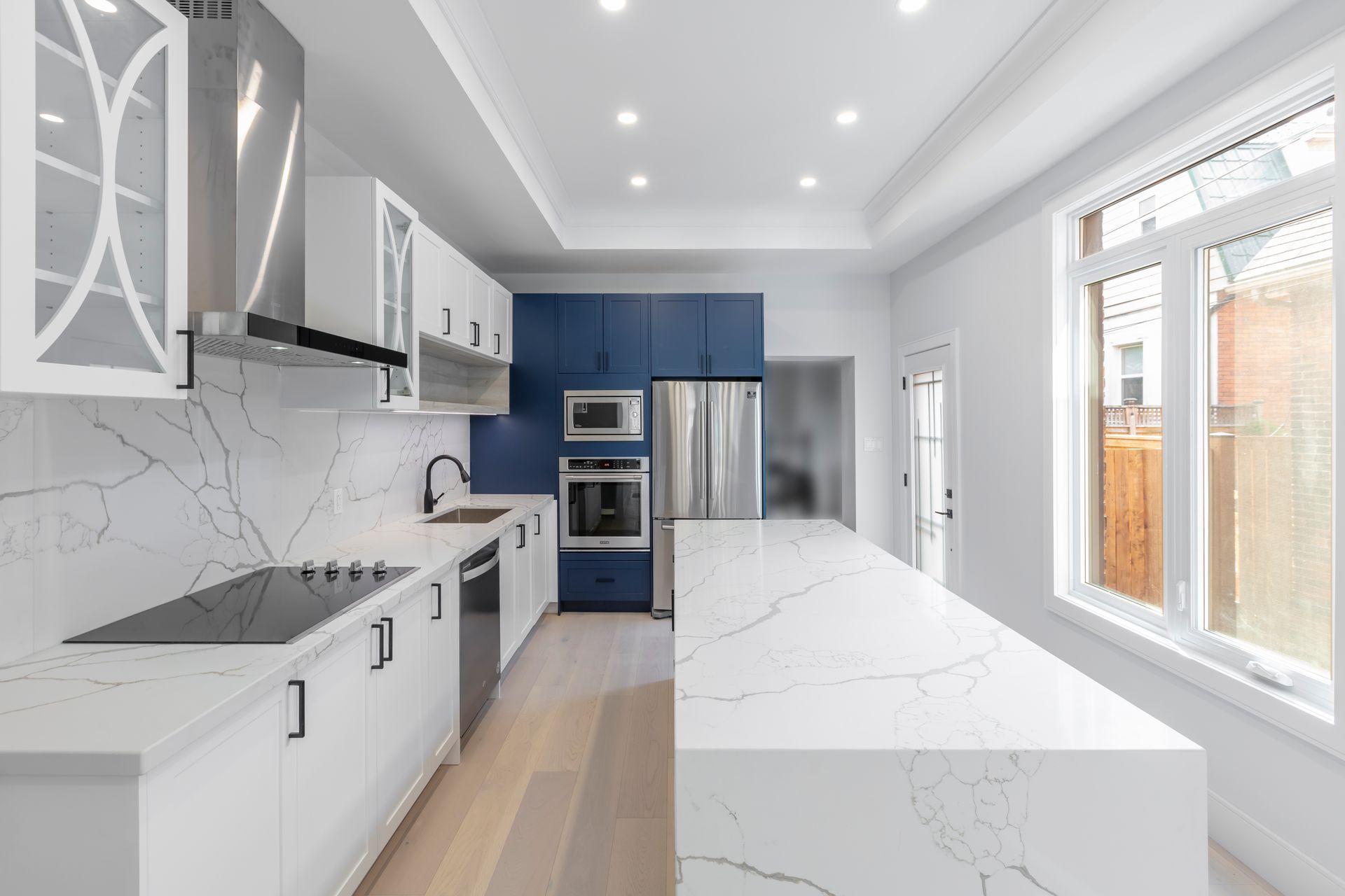 Modern white kitchen with marble island, blue cabinets, built-in oven, and large windows