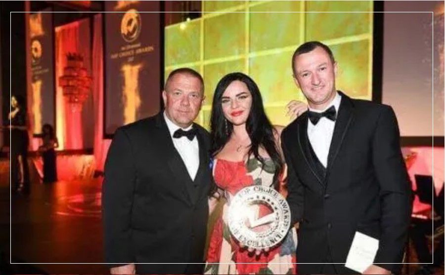 A woman in a red dress is standing between two men in tuxedos holding a trophy.