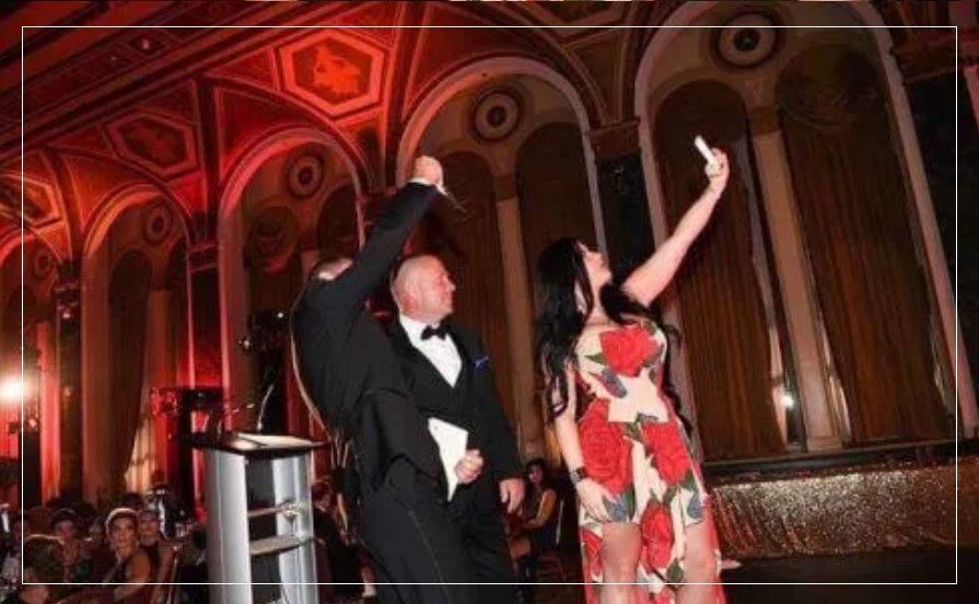 A man in a tuxedo and a woman in a dress are taking a selfie in a ballroom.