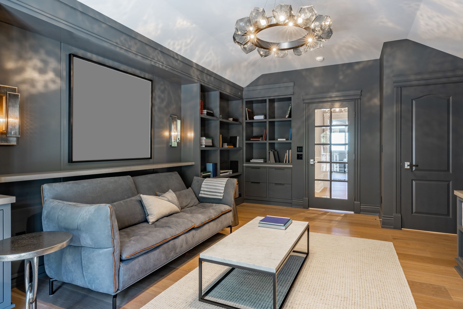 Dark gray study with a sofa, bookshelves, and a glass-paned door leading to another room; a modern chandelier hangs from the vaulted ceiling.
