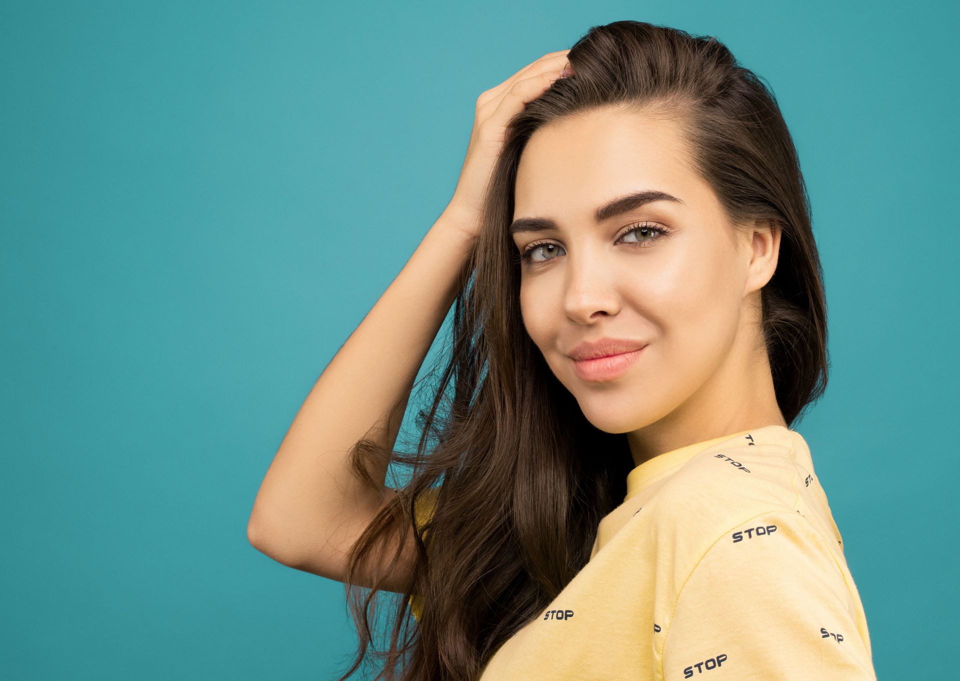 Woman posing against a teal background, hand on head, wearing a yellow shirt, smiling softly