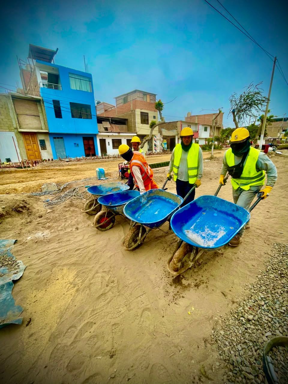 Obreros empujando carretillas azules a través de una obra en construcción embarrada bajo un cielo nublado.