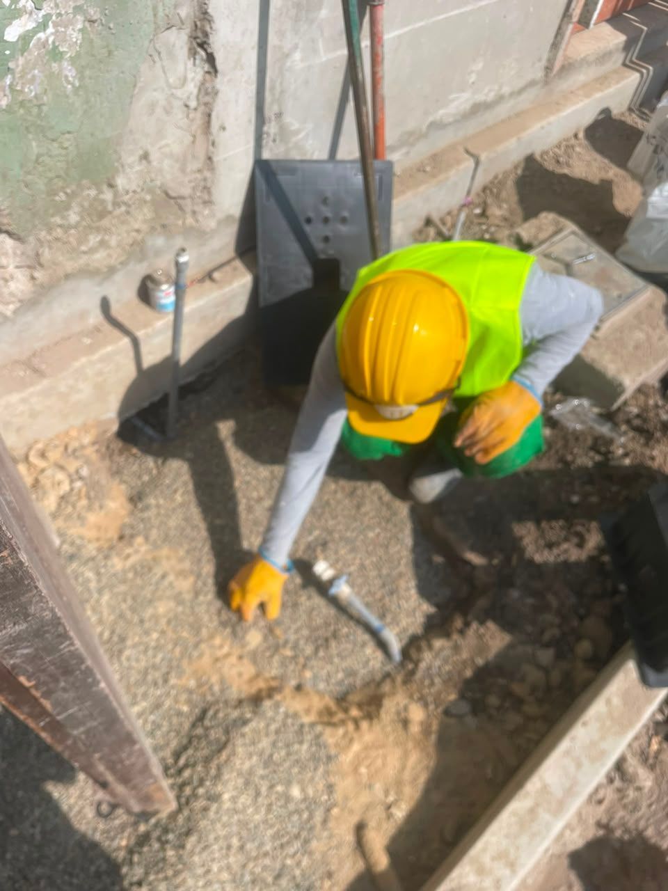 Obrero con casco y chaleco amarillos utilizando una herramienta en una zanja de hormigón en una obra de construcción.