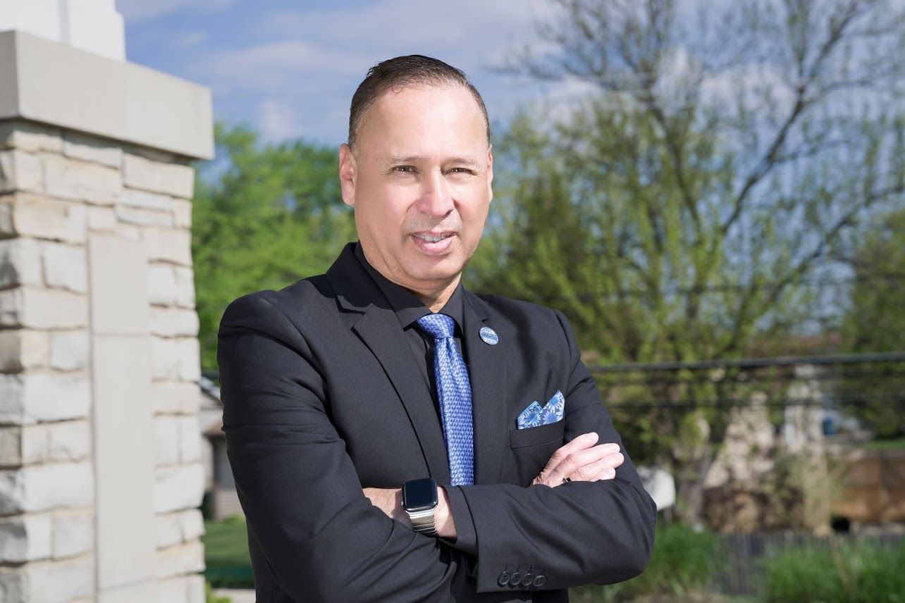 Man in black suit with arms crossed, standing outdoors by a stone wall and trees on a sunny day