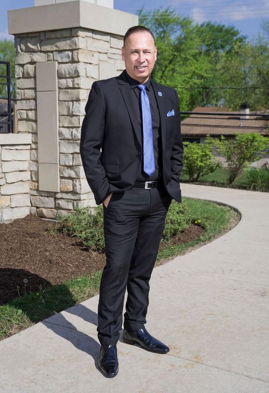 Man in a black suit stands on a curved sidewalk beside a stone pillar and greenery.