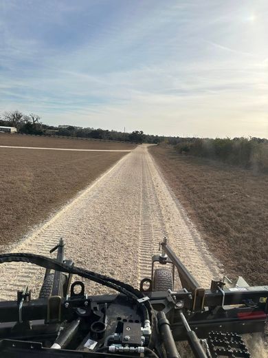 View from a farm machine driving down a gravel path between plowed fields under a pale sky