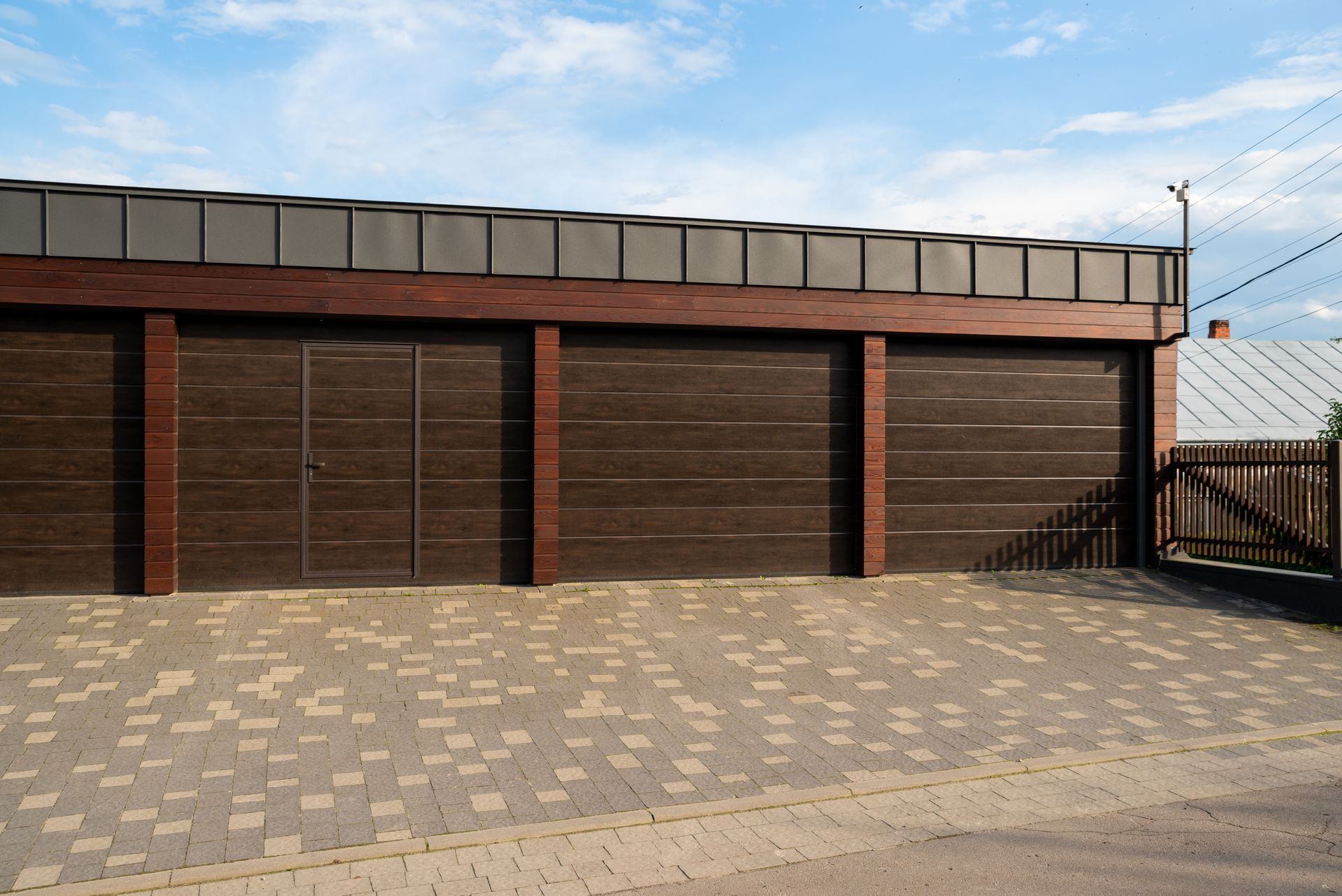 Brown garage with three doors under a brown roof and a paved driveway, with a fence and a house in the background.