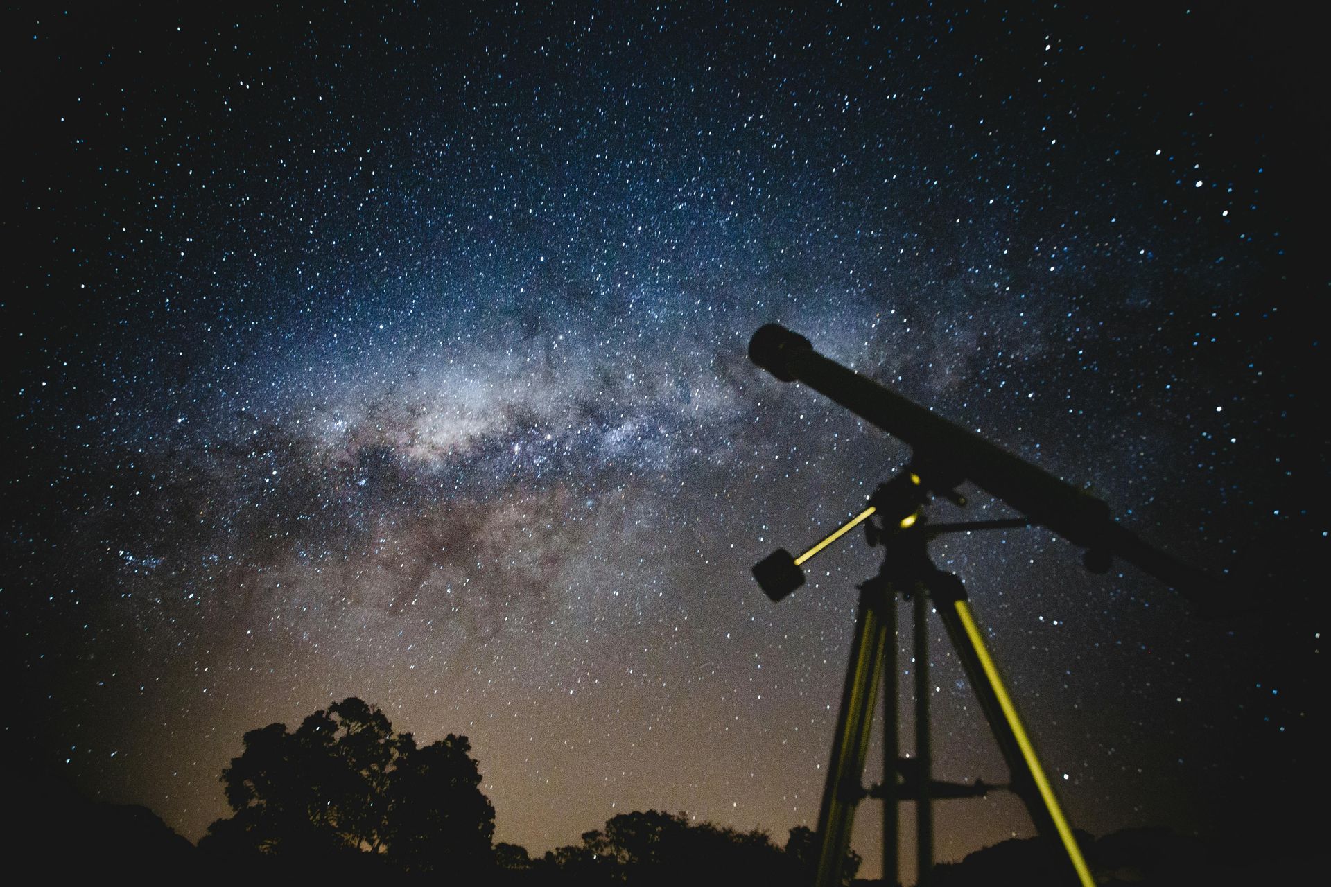 A silhouette of a telescope on a tripod pointed toward a vast, star-filled night sky and the Milky Way.