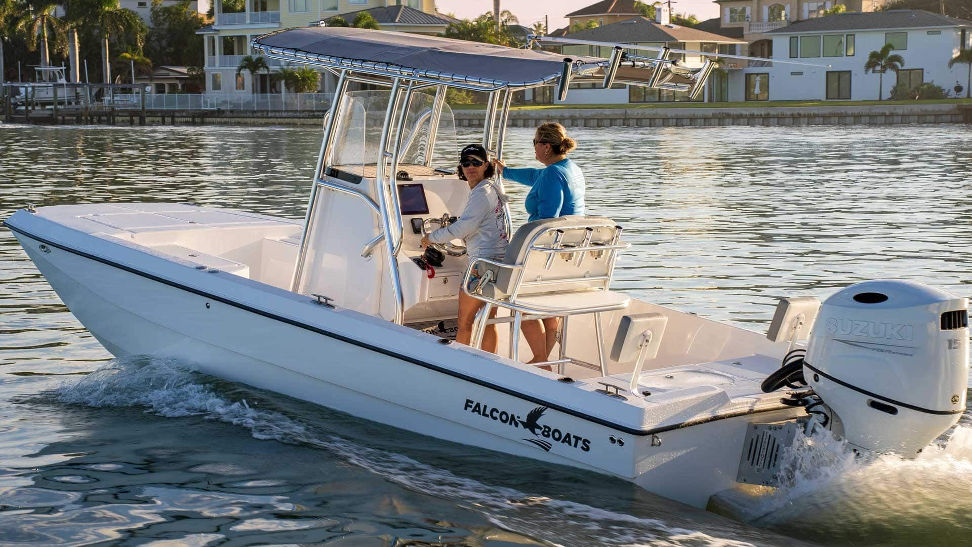 Two women piloting a white center-console boat on a sunny waterway, residential buildings in the background.