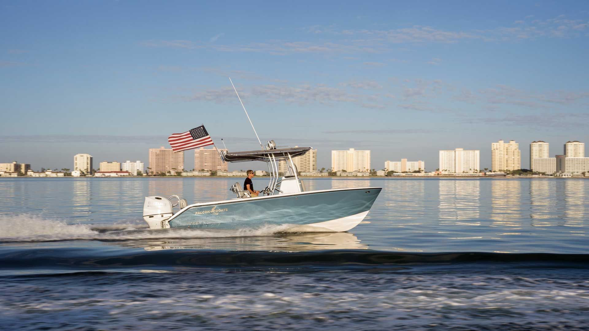 A blue and white motorboat with an American flag speeds across calm water toward a cityscape on a sunny day.