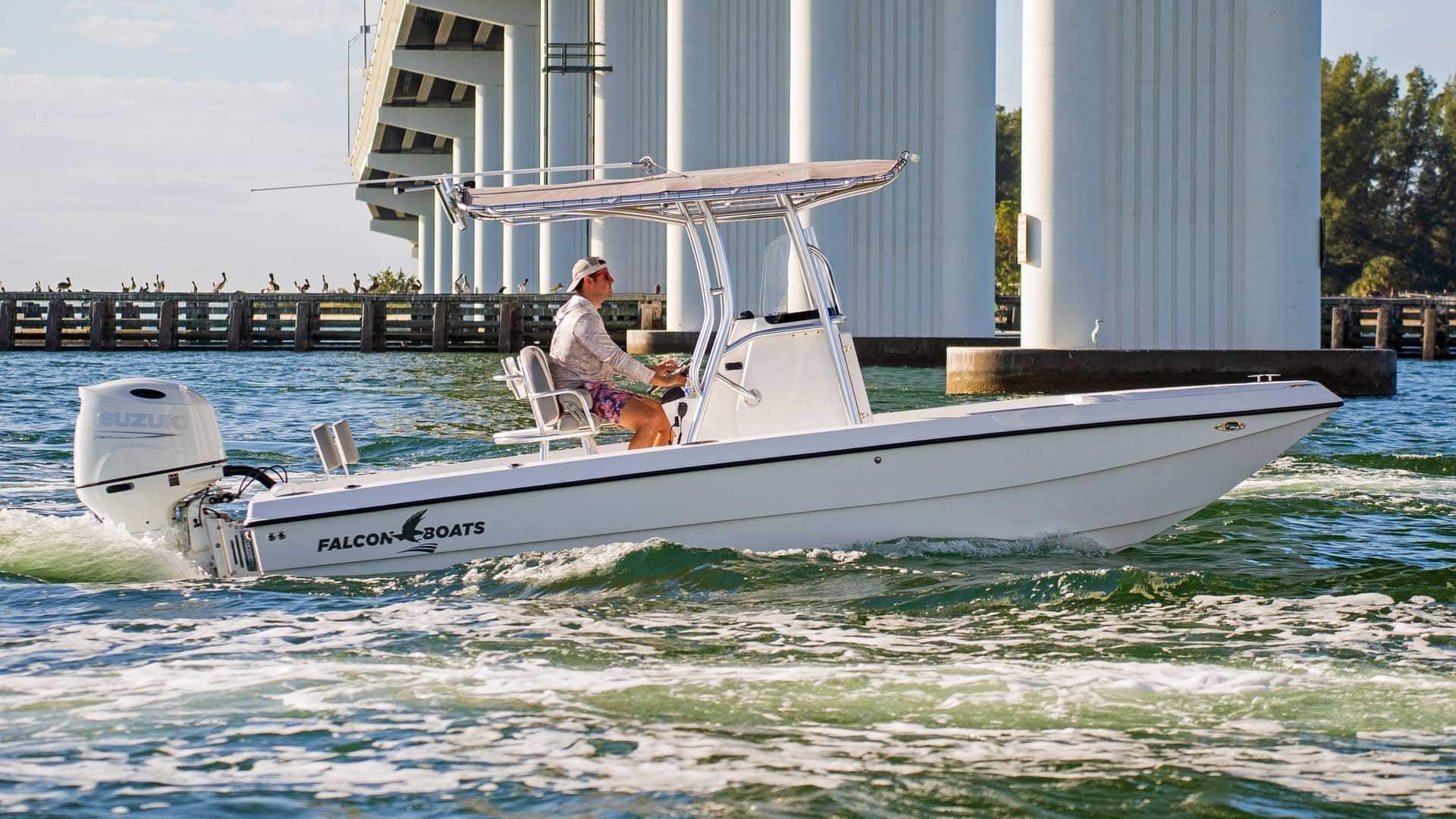 White boat with a person at the helm traveling under a bridge. The boat is on the water, and the sky is blue.