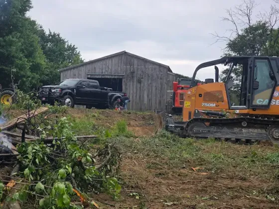 Excavator and log splitter beside a barn, with a black pickup truck and brush pile in a muddy yard