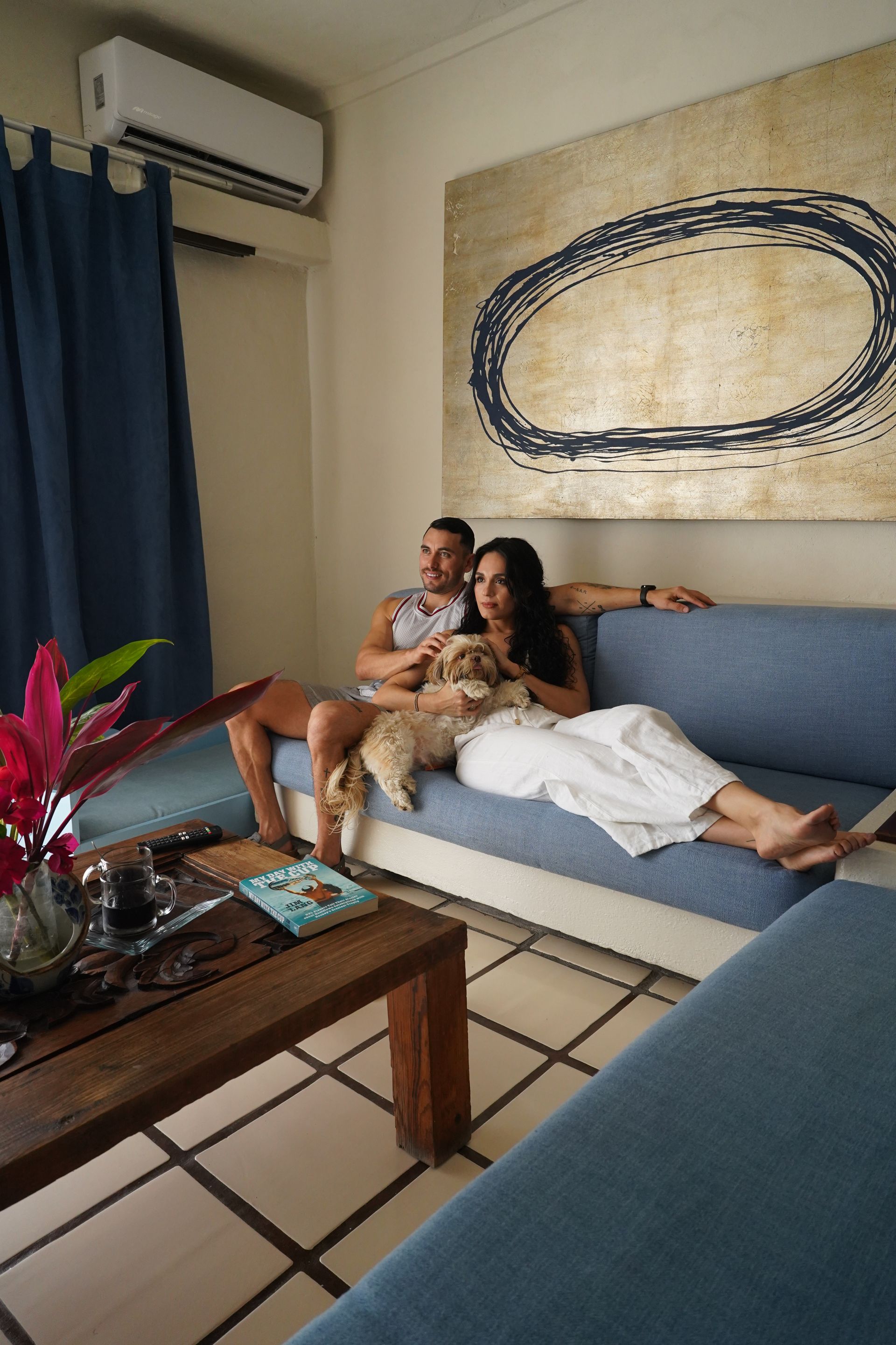 Couple lounging on a blue sofa in a living room with a coffee table, curtain, and wall art.