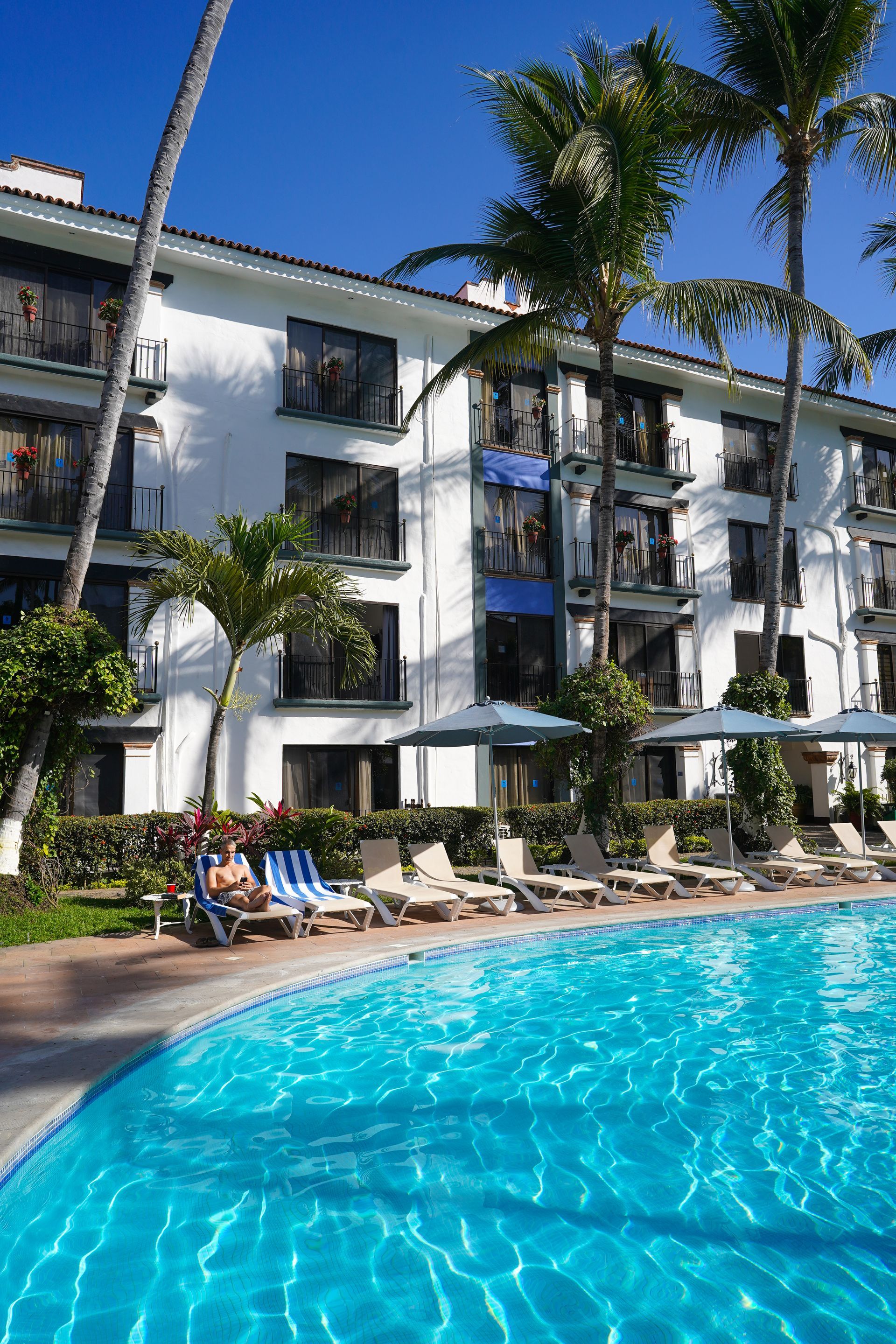 Hotel with swimming pool and lounge chairs; palm trees and blue sky.