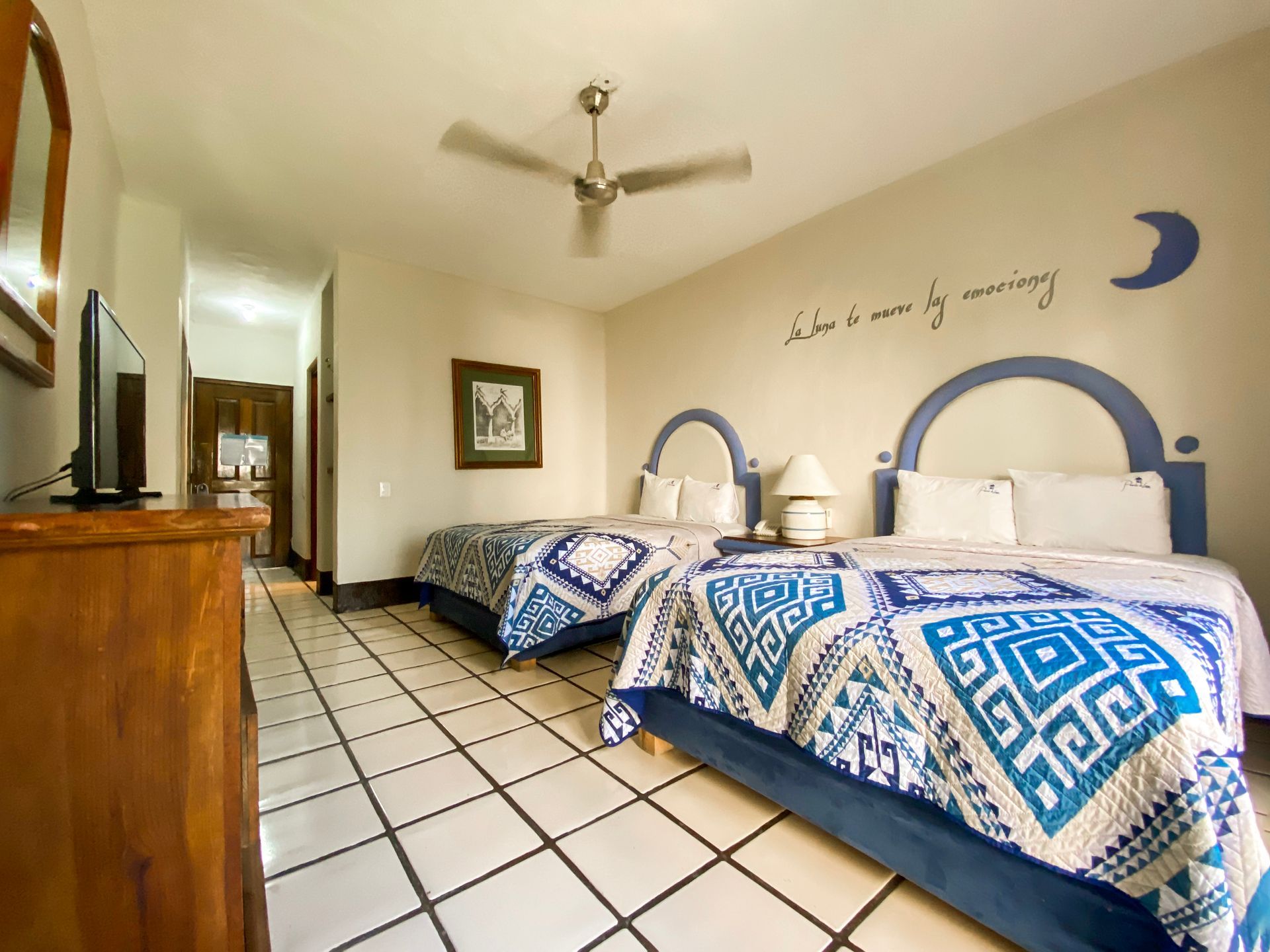 Two beds with blue and white quilts in a bedroom; a lamp and artwork hang on the walls.