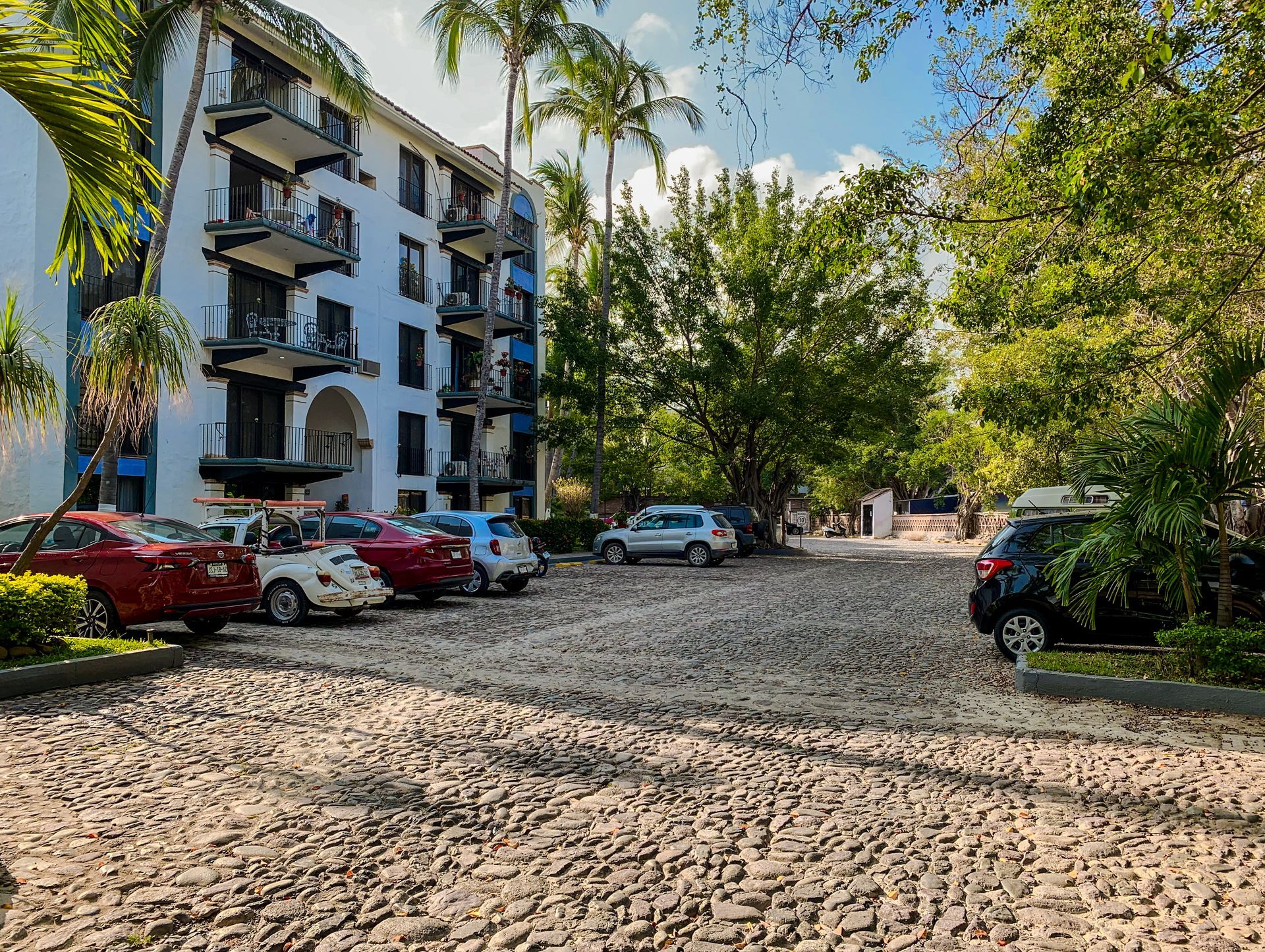 Apartment courtyard with parked cars, palm trees, and a cobblestone drive under bright sunlight