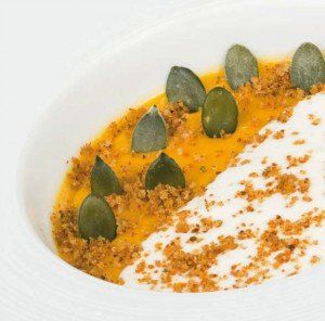 Purea d'arancia con foglie di erbe aromatiche verdi e spuma bianca servita in una ciotola bianca minimalista.