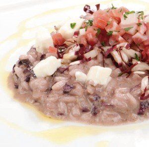 Risotto cremoso ai funghi con pomodori a cubetti, erbe aromatiche e scaglie di formaggio, servito su un piatto bianco.