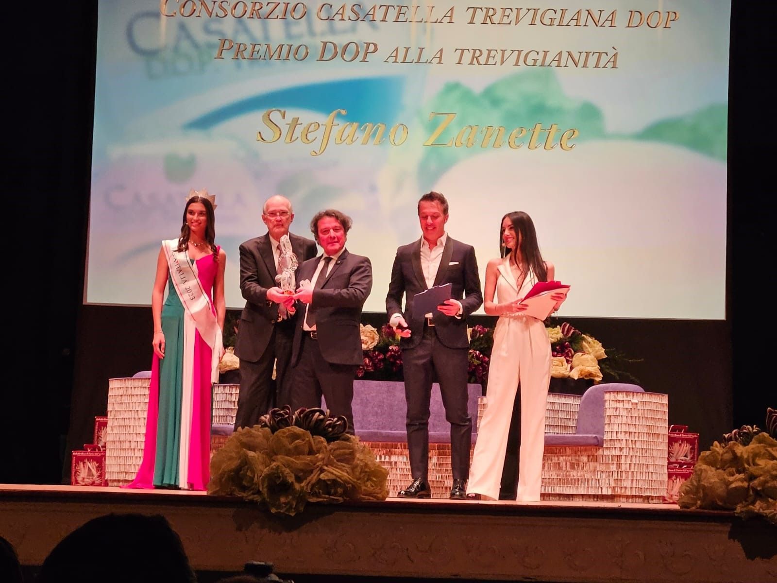 Cinque persone sul palco che reggono i premi davanti a uno sfondo dell'evento proiettato