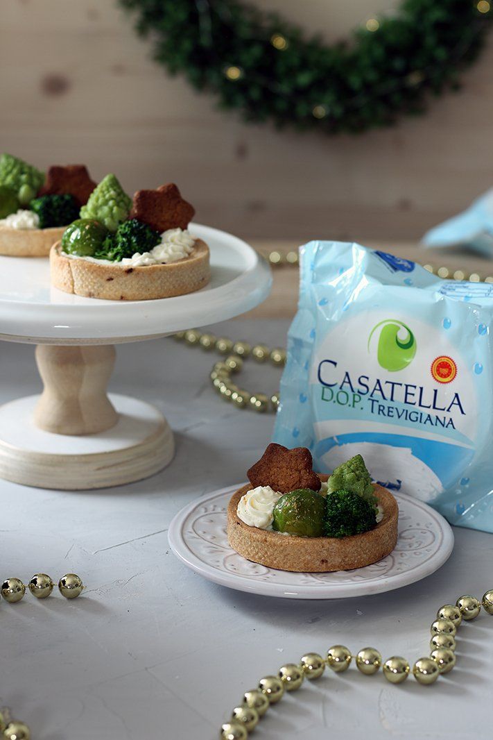 Esposizione di dolci natalizi con mini crostatine e confezione blu di Casatella su un tavolo bianco.