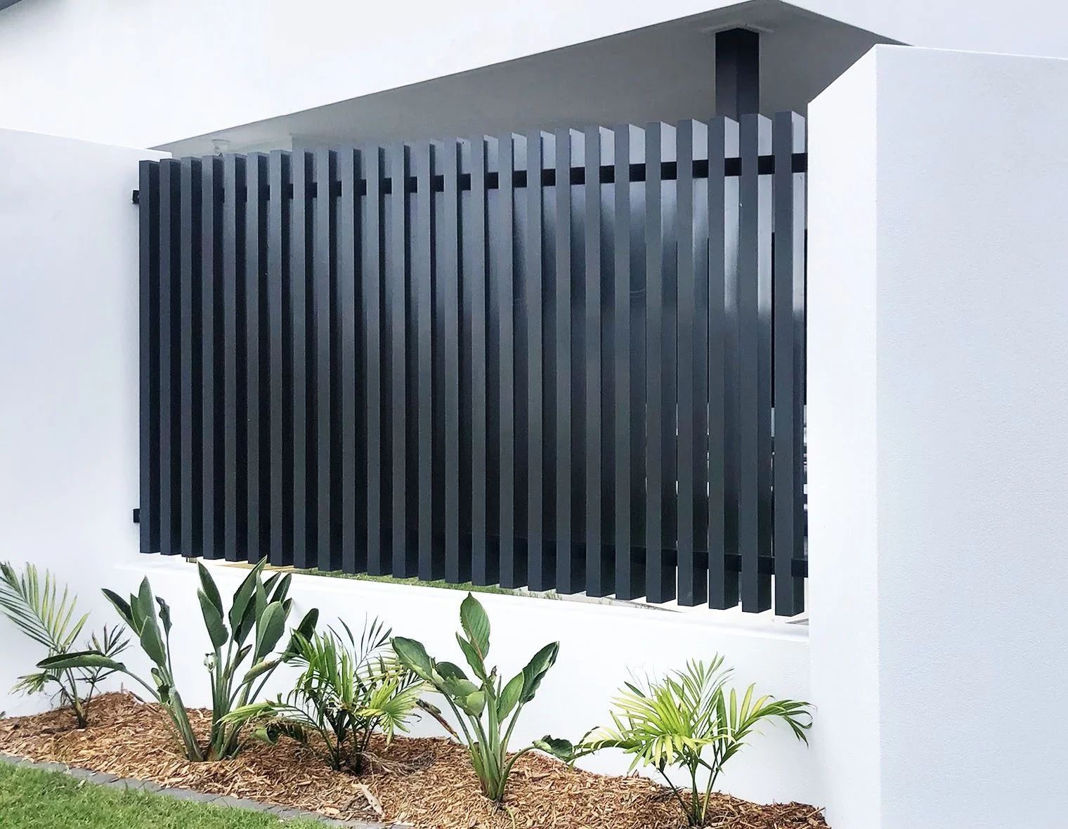 A black aluminium batten fence is sitting on a white wall next to a garden.
