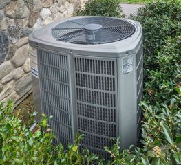 Outdoor air conditioner unit beside a stone wall, surrounded by green shrubs