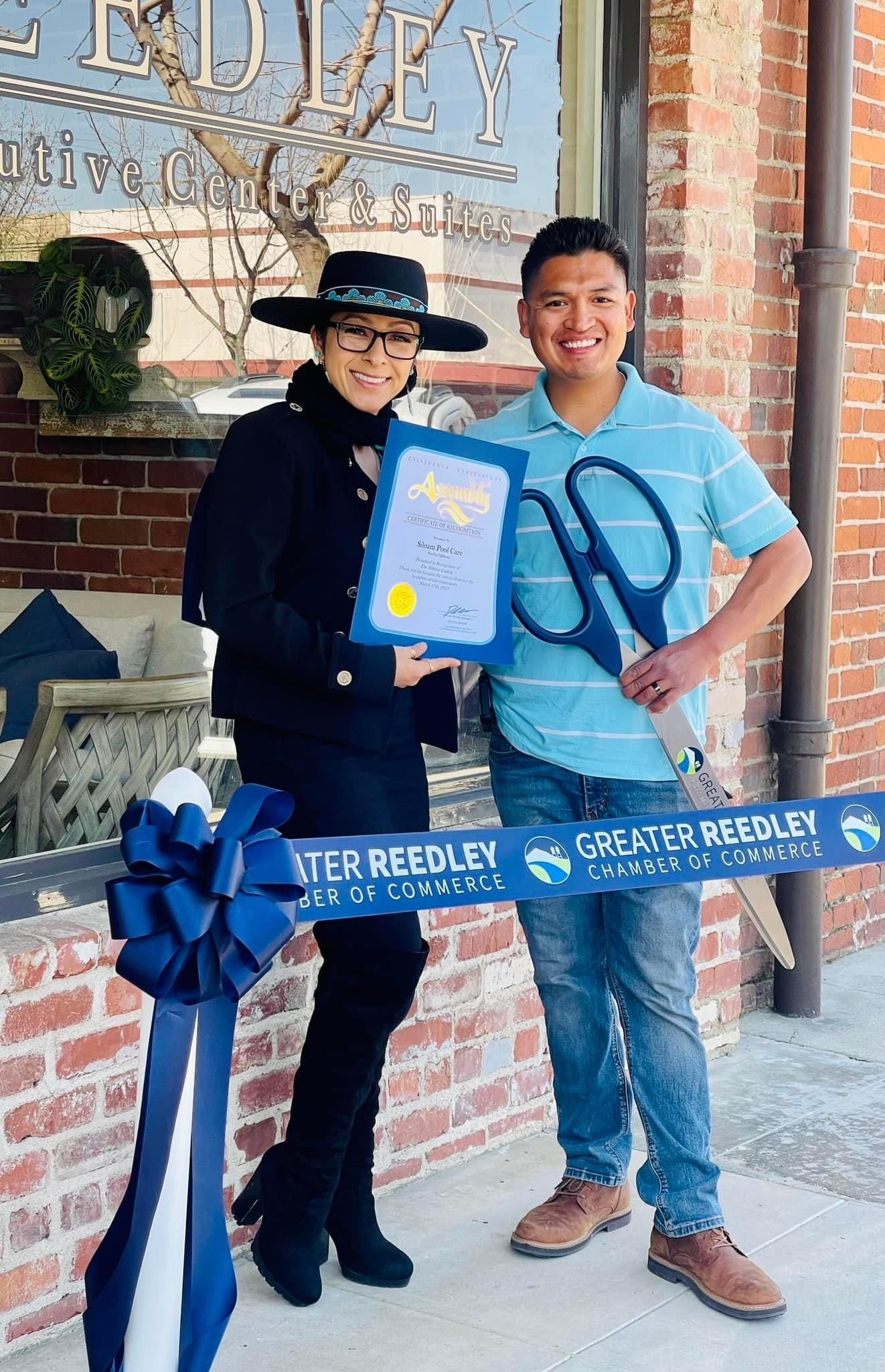 Ribbon-cutting ceremony, holding a certificate and oversized scissors, with blue ribbon.